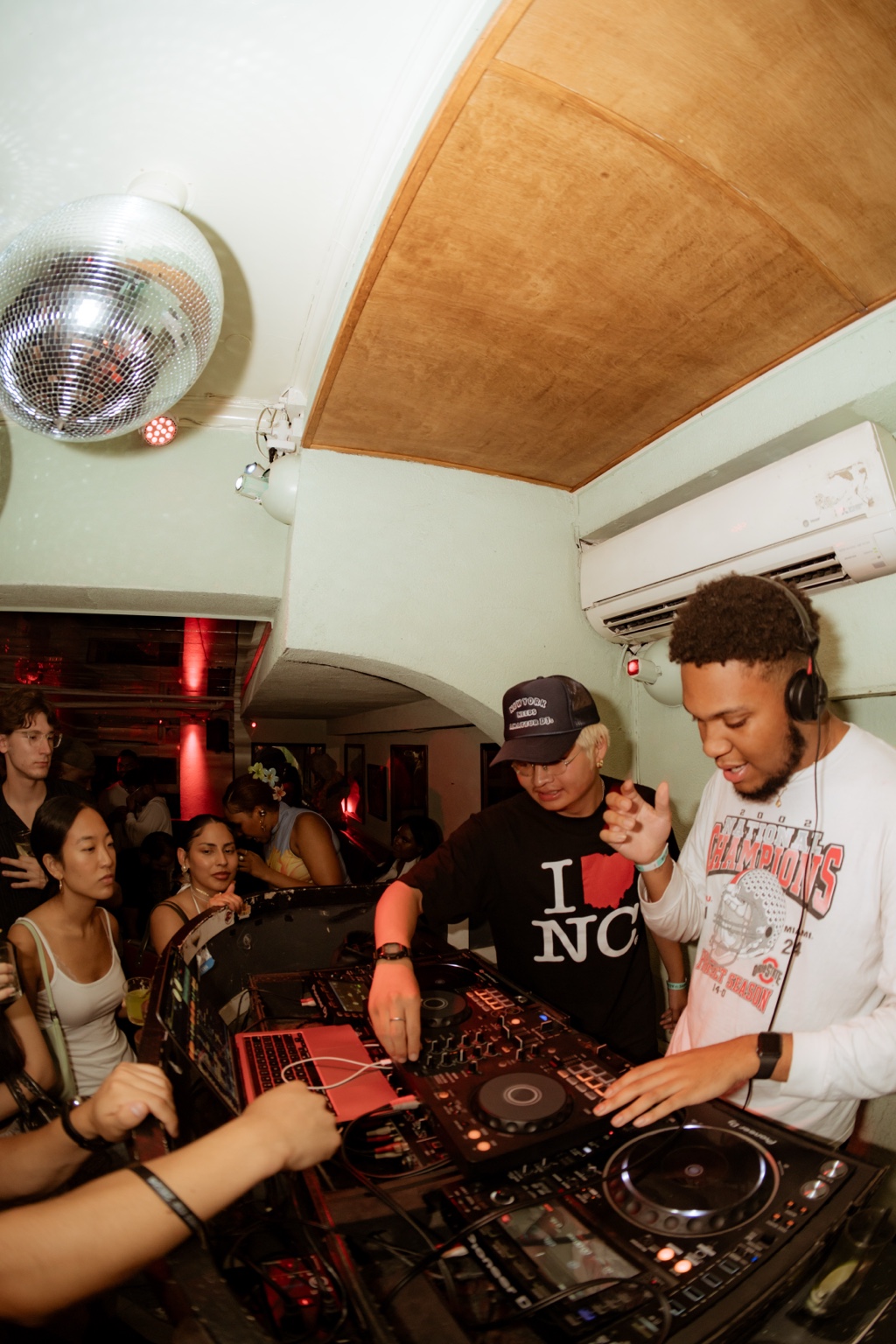 A crowded DJ booth beneath a disco ball
